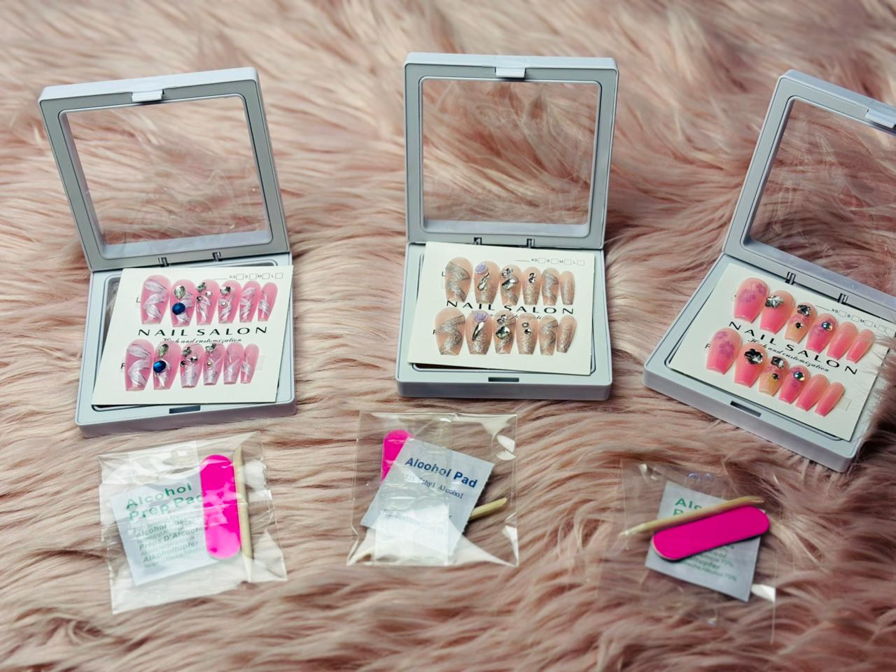 Assorted ready-to-wear press-on nail sets displayed in compact storage cases on a soft fur background, including pink and nude designs with rhinestones, along with nail glue tabs, alcohol pads, and mini nail file for easy at-home application.