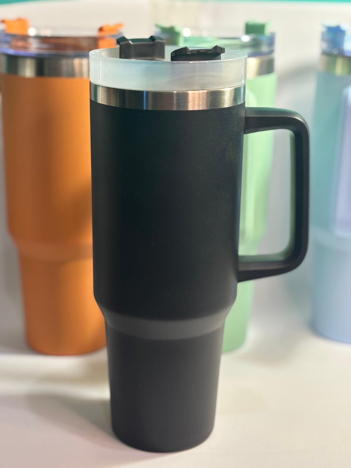 Black insulated mug with a handle on a white surface, surrounded by other colorful mugs.