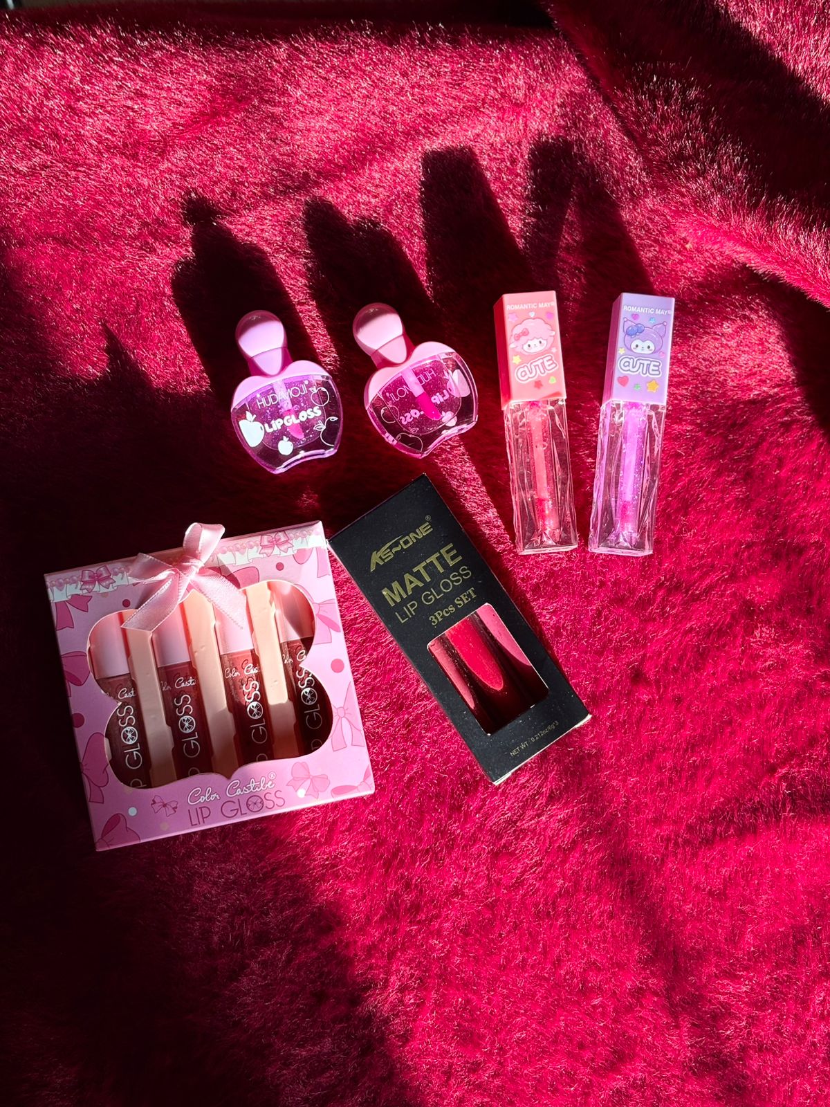 A collection of assorted lip gloss sets including heart-shaped glosses, cartoon-tube glosses, a pink gift box gloss set, and a matte lip gloss trio, all arranged on a bright red furry background.