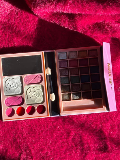 An opened BLs Revlon makeup palette showing blush pans, lip colors, and multiple eyeshadows displayed on a red textured surface.