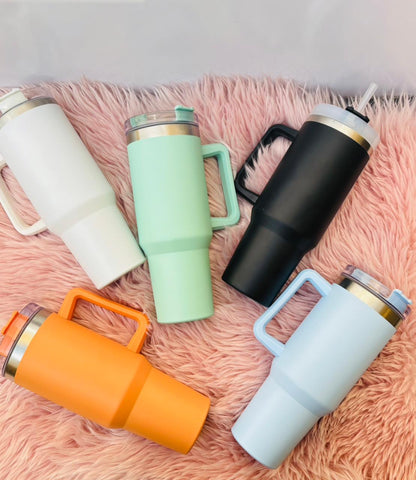 Aesthetic display of six matte stainless steel insulated tumbler cups with straws and handles in pastel and bold colors arranged on a soft pink background.