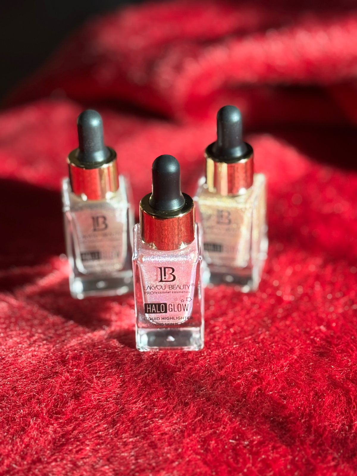 Three luxury liquid highlighter serums with dropper caps displayed on a textured red fabric surface."