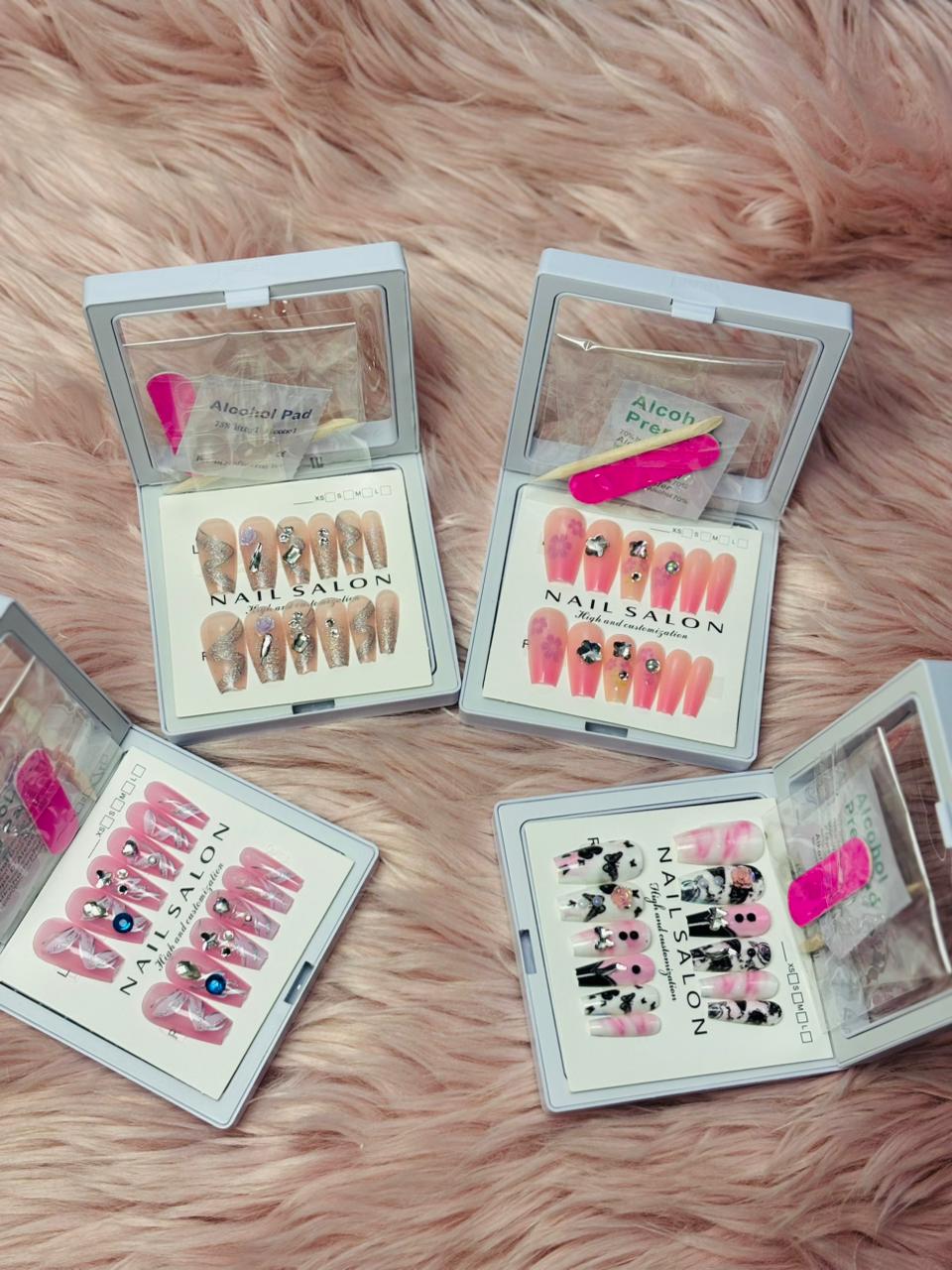 Assorted luxury press-on nail sets displayed in transparent storage cases on a soft fur background, including nude, pink, and printed nail designs with rhinestones, alcohol prep pads, and nail tools for easy application.