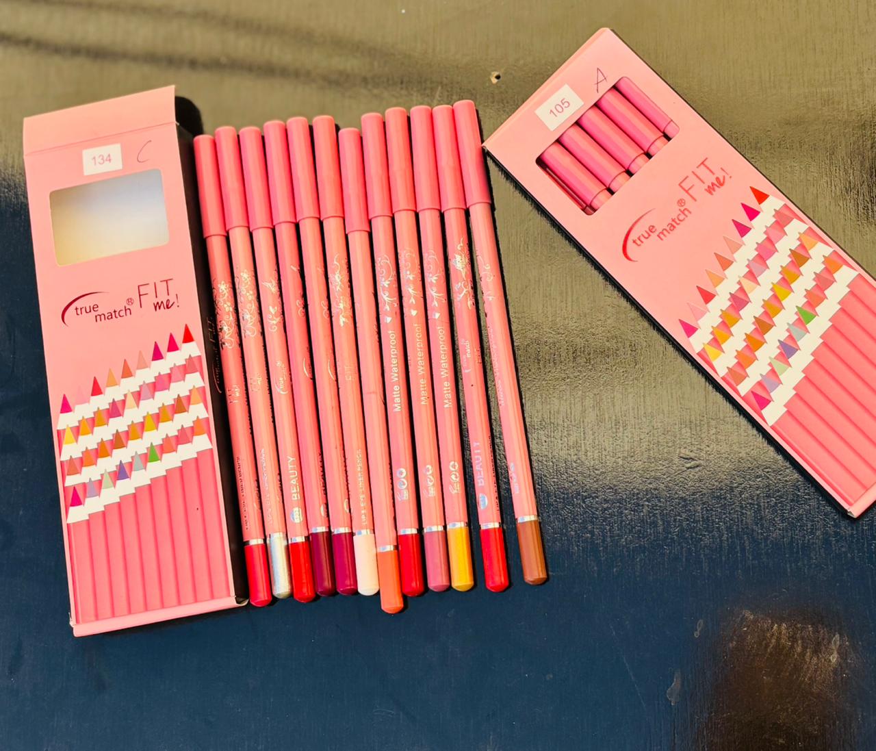 A row of colorful matte waterproof lip and eye pencils laid out on a dark surface, revealing various shades from nude to bold reds and oranges.