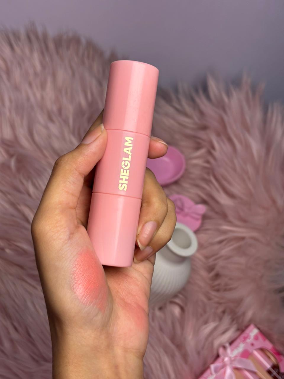 SHEGLAM soft pink liquid blush held in a hand beside a small decorative vase on a pink surface.